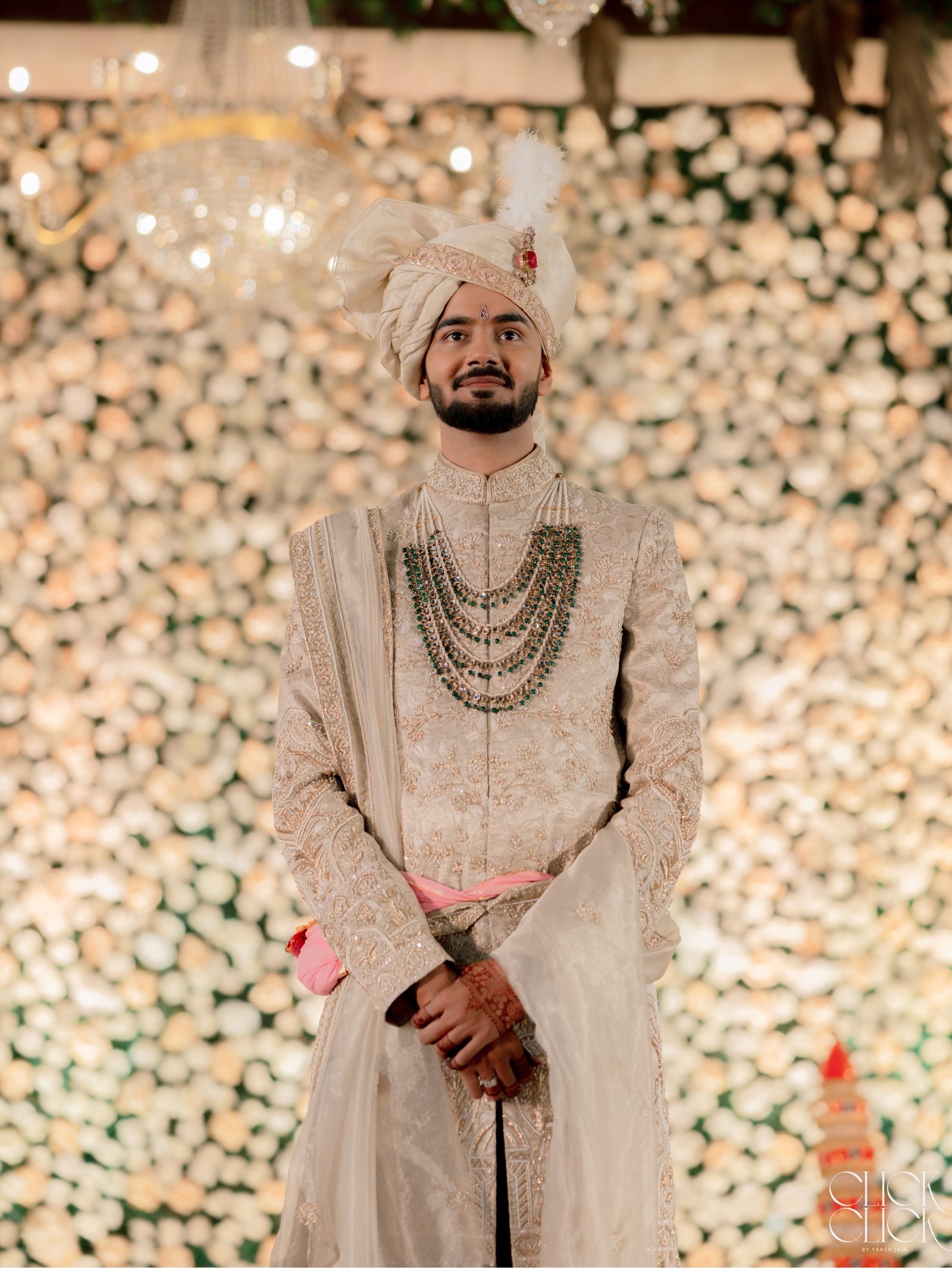 A groom in traditional wedding attire before a wall of flowers